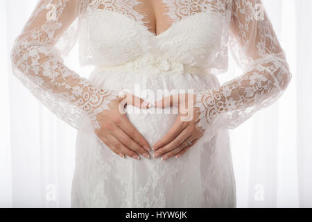 Les mains d'une femme qui se tient en forme de cœur sur son ventre enceinte. Elle est vêtue d'une robe de maternité en dentelle. Banque D'Images