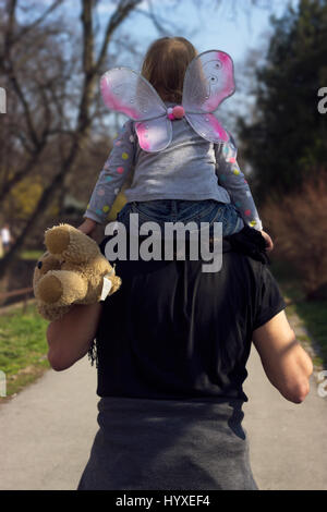 Père exerçant peu bébé fille, avec des ailes de papillon et ours en peluche, sur ses épaules Banque D'Images