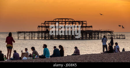 Des foules de gens sont silhouetté contre un rouge lumineux ciel coucher soleil sur la plage de Brighton, avec les ruines de la jetée Ouest Comité permanent sur la mer derrière. Banque D'Images