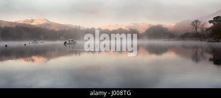 La brume s'élève des eaux calmes du lac Windermere, à Ambleside Pier, à côté de l'automne et forestiers dans le cadre de montagnes enneigées en Angleterre du Lake District. Banque D'Images