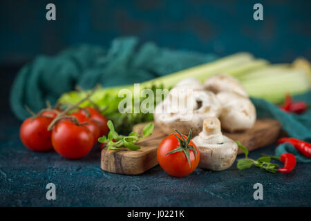 Champignons Champignons frais sur une planche à découper en bois verts, persil, céleri, tomates cerise et piment rouge chaud sur un bleu foncé backgrou Banque D'Images