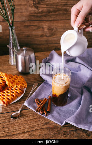 La main de la femme avec la coulée de verre de lait crème avec du café. Les bâtons de cannelle, biscuits maison et bouquet de fleurs sur un fond de bois. Le petit-déjeuner gratuit. Banque D'Images