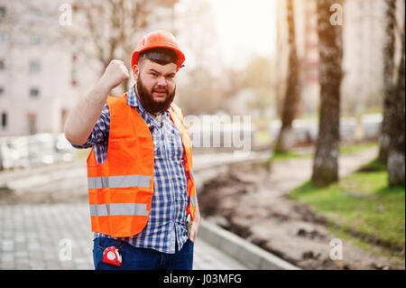 Portrait de l'homme travailleur barbe en colère brutale construction worker in costume orange sécurité casque contre trottoir avec montrant des bras. Banque D'Images