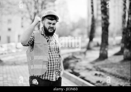 Portrait de l'homme travailleur barbe en colère brutale construction worker in costume orange sécurité casque contre trottoir avec montrant des bras. Banque D'Images