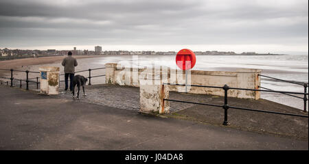 Un homme marche d'un grand chien sous un ciel gris sur la promenade de Saltcoats sur la côte d'Ayrshire. Banque D'Images