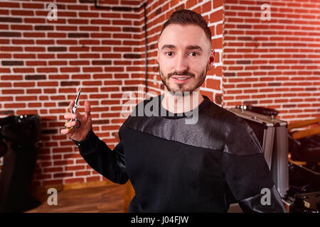 Beau coiffure barbu elégante en noir l'usure est la tenue d'une paire de ciseaux tout en se tenant à la coiffure. Banque D'Images