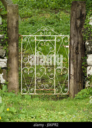 Ancien, peint en blanc, orné, porte de jardin en fer forgé avec tourbillons entre poteaux de bois avec l'herbe verte. Banque D'Images