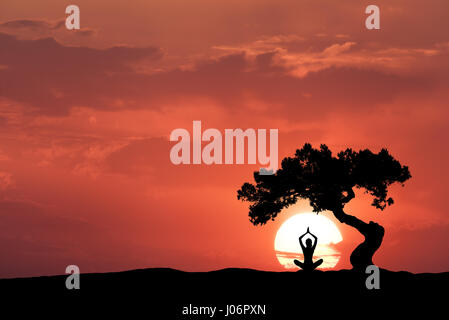 Silhouette d'un sitting woman practicing yoga avec les armes sous l'arbre de travers sur la colline en arrière-plan de soleil et ciel rouge couleur wit Banque D'Images