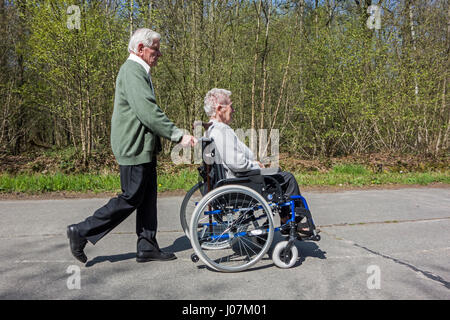 Mari à la retraite âgées handicapées prendre épouse dans fauteuil roulant pour une promenade dans le parc lors d'une journée ensoleillée au printemps Banque D'Images