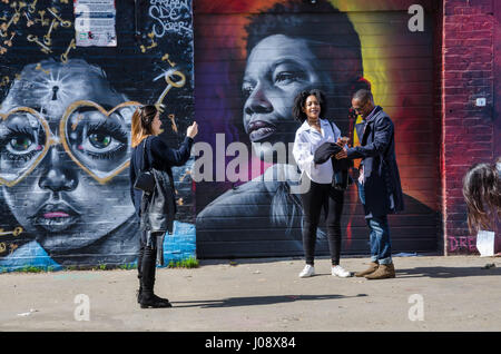 Les touristes d'admirer et de poser pour des photographies avec le street sont coloré et dynamique qui couvre les murs d'une aire de stationnement au large de Brick Lane à Londres. Banque D'Images