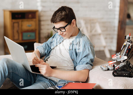 Être attentif. Garçon intelligent attrayant en gardant un ordinateur portable sur la jambe appuyée sur le canapé et smiling Banque D'Images