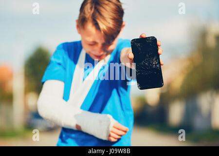 Petit garçon avec fracture de la main blessé après accident montrant téléphone mobile endommagé Banque D'Images