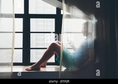 Homme triste solitaire assis derrière le rideau avec un téléphone mobile à la fenêtre pendant les jours sombres. Banque D'Images