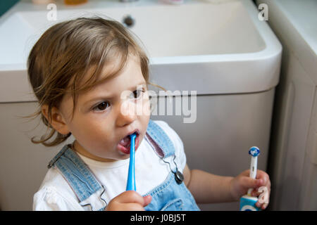 Tout-petit fille se brosser les dents à l'aide d'une brosse à dents pour enfants, adultes et de tenir une brosse à dents électrique dans son autre main. Elle est de 16 mois Banque D'Images