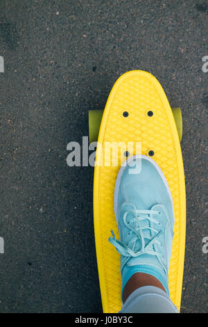 La jambe de la femme en bleu jeans et baskets jaune debout sur la planche à roulettes, de point de vue, cadrage vertical Banque D'Images