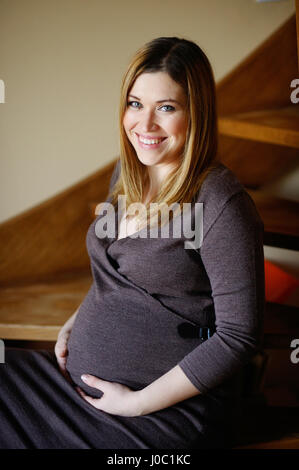 Belle jeune femme enceinte est assise sur les marches d'une échelle en bois. Elle embrasse les mains soigneusement l'estomac. Femme regarde dans la caméra et sourit. Banque D'Images