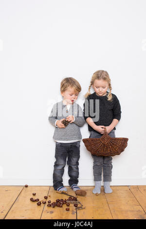 Jeune fille et garçon, girl holding panier en osier, boy holding pine cone, pommes de pin et conkers sur plancher Banque D'Images