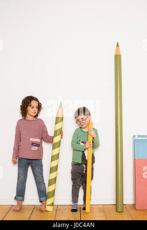 Jeune fille et un garçon debout, tenant giant size crayons, papeterie géante leaning on wall à côté de lui Banque D'Images