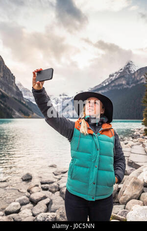 Femme debout au bord du lac, en tenant avec selfies smartphone, Lake Louise, Alberta, Canada Banque D'Images