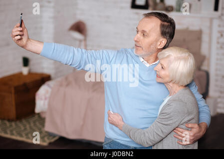 Nous sommes modernes. Portrait of handsome man embrassant sa femme et en tenant debout selfies à la maison. Banque D'Images