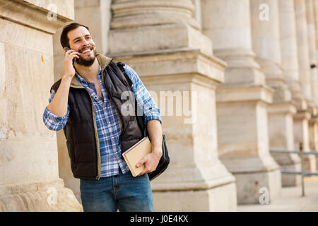 Smiling Young man with books et son sac à dos à l'aide d'un téléphone portable à l'avant et à l'ancien bâtiment classique avec grandes colonnes Banque D'Images