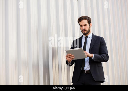 Homme d'affaires moderne et élégant en costume et à l'aide d'une tablette pour son travail quelque part dehors à côté d'un mur nu gris Banque D'Images