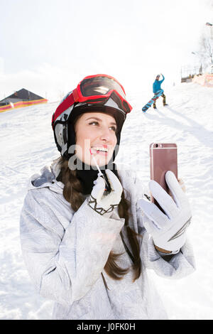 Smiling young female snowboarder appliquant un brillant à lèvres tout en holding smartphone Banque D'Images