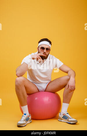 Image of young sportsman wearing glasses sitting on fitness ball isolé sur fond jaune. Looking at camera holding cigarette. Banque D'Images