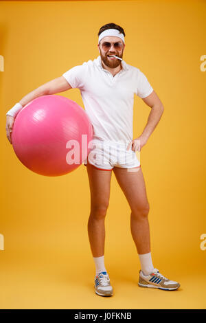 Image de beau jeune sportsman wearing glasses holding permanent isolé fitness ball sur fond jaune. Looking at camera holding cigarette. Banque D'Images