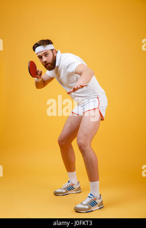 Photo de happy young sportsman habillé en blanc T-shirt isolés sur fond jaune holding racket de tennis de table. En regardant la caméra. Banque D'Images
