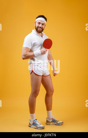 Image of young sportsman habillé en blanc T-shirt isolés sur fond jaune holding racket de tennis de table. En regardant la caméra. Banque D'Images