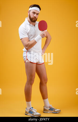 Photo de beau jeune sportsman habillé en blanc T-shirt isolés sur fond jaune holding racket de tennis de table. Regardant est venu Banque D'Images