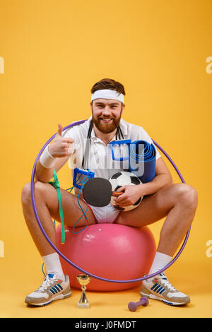 Portrait of a happy sports homme assis sur un ballon de fitness et showing Thumbs up geste isolé sur un fond orange Banque D'Images