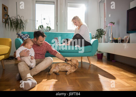 Père assis sur le plancher et avec bébé dans ses bras jouer avec chien femme assis sur le canapé et de faire les achats en ligne par carte de crédit Banque D'Images