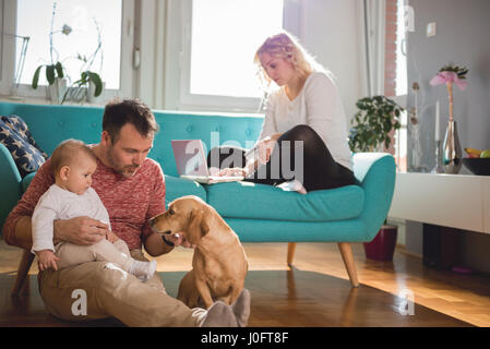 Père assis sur le plancher et avec bébé dans ses bras jouer avec chien femme assis sur le canapé et de faire les achats en ligne par carte de crédit Banque D'Images