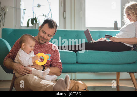 Père assis sur le plancher et la tenue de bébé dans ses bras tandis que femme assis sur le canapé et de faire les achats en ligne par carte de crédit Banque D'Images