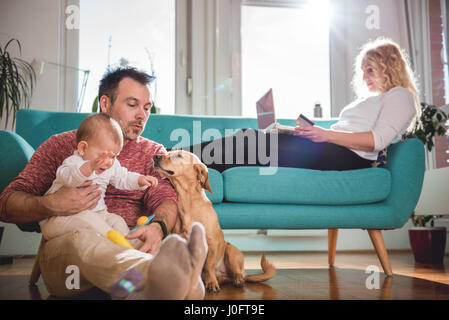 Père assis sur le plancher et avec bébé dans ses bras jouer avec chien femme assis sur le canapé et de faire les achats en ligne par carte de crédit Banque D'Images