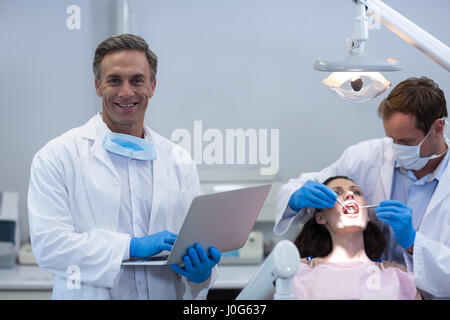 Portrait de dentiste holding laptop while collègue examinant une patiente à la clinique dentaire Banque D'Images