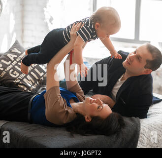 Les jeunes parents avec petit bébé s'allonger sur lit et jetez-le en place. Ils rient de plaisir. Banque D'Images