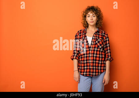 Jolie femme bouclés dans une chemise à carreaux et jeans debout sur un fond orange avec l'espace vide pour le texte à côté d'elle Banque D'Images