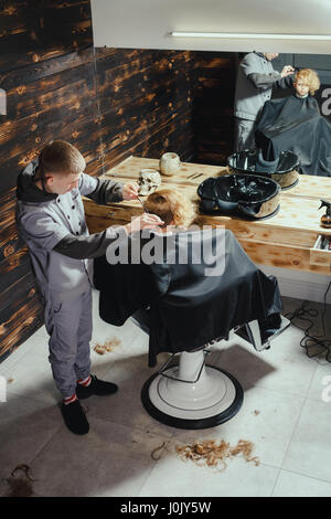 Petit garçon se coupe par coiffure While Sitting in Chair At Barbershop. Thème Barbershop Banque D'Images