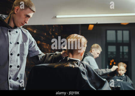 Petit garçon se coupe par coiffure While Sitting in Chair At Barbershop. Thème Barbershop Banque D'Images