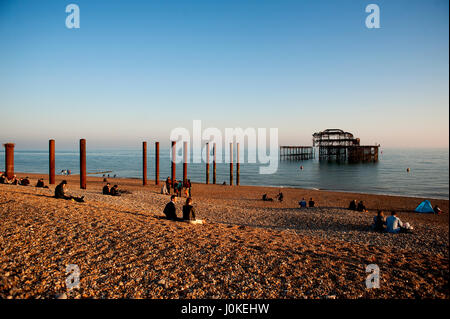 Les gens s'assoient sur la plage de galets à Brighton West Pier un célèbre monument sur la côte sud de l'Angleterre, maintenant un squelette rouille brûlée. Banque D'Images