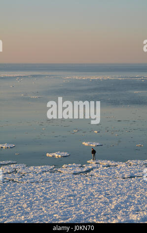 Un homme se tient sur le bord d'une plaque de glace et regarde vers le bas dans l'eau. L'immensité du lac Michigan s'étend au-delà de lui. Banque D'Images