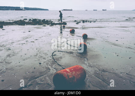 Femme debout à proximité de rochers à marée basse avec des flotteurs qui serpente le long de la chaîne du sable mouillé. Banque D'Images
