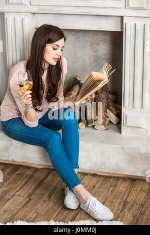 Jeune femme séduisante assis près de cheminée reading book et de boire le vin blanc Banque D'Images