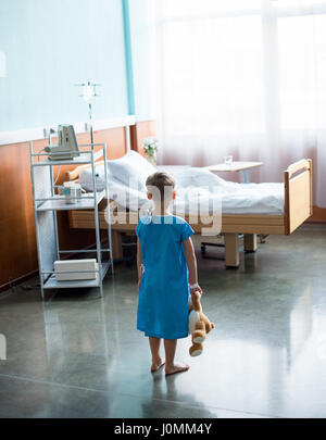 Vue arrière de petit garçon aux pieds nus avec teddy bear standing in hospital room Banque D'Images