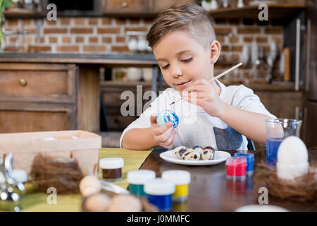 Cute smiling little boy painting easter egg dans la cuisine Banque D'Images