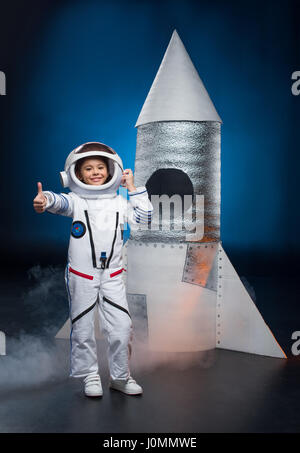 Smiling little girl en costume d'astronaute debout près de lancement de roquettes et de showing thumb up Banque D'Images