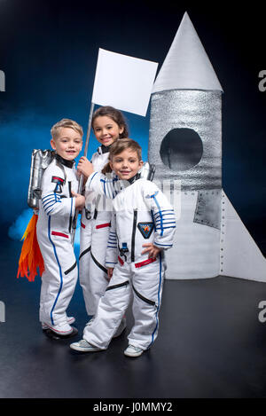 Deux petits garçons et filles dans l'espace suits holding flag près de rocket and smiling at camera Banque D'Images